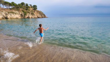 Çocuk deniz kıyısına doğru koşuyor su sıçrıyor kristal berrak turkuaz sulara dalmak için Costa Brava, Girona, İspanya 'da arka planda bir uçurum var.