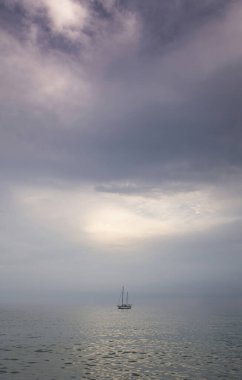 Gün batımında tamamen sakin bir şekilde denizin ortasına demir atmış küçük bir yelkenli. Leylak renginde bulutlu bir gökyüzü, sakinlik, izolasyon, minimalizm ve yalnızlık hissi, dikey kopyalamak için yer.