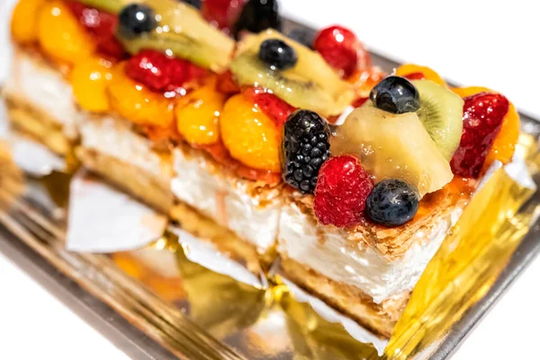 close up of a puff pastry and cream cake with red wild berries, kiwi fruit pieces, pineapple, mandarin slices on a white table, selective focus on one blackberry, horizontal