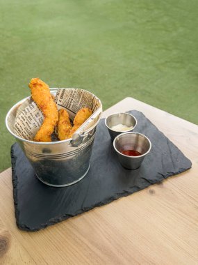 Metal bir kovanın içinde tavuk parmakları ve tahta bir masanın üzerinde farklı soslar bulunan iki metal kap. Arka planda dikey olmayan çimenler.