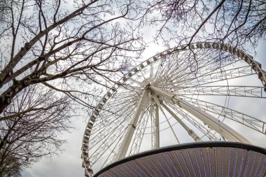 Bulutlu bir gökyüzüne karşı çıplak ağaç dalları tarafından çerçevelenmiş geniş bir dönme dolabın alçak açılı görüntüsü. Kışın şehir eğlence merkezi, eğlence, turizm ve şehir eğlencesi sembolü, yatay