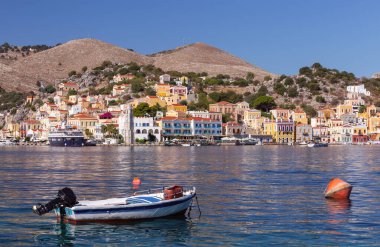 Yunanistan 'ın Symi kentindeki Symi adasının limanı boyunca uzanan geleneksel mimari, sakin deniz kıyısında renkli binalar ve teknelerle.