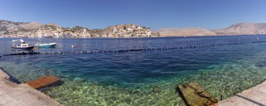 Yunanistan 'ın Suriye kentindeki Symi adasının geniş panoramik manzarası, turkuaz su, balıkçı tekneleri ve renkli binalı kıyı tepelerini gösteriyor.