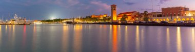 Aydınlatılmış Rodos limanı, Yunanistan günbatımında demirli yatlar, Büyük Üstad Sarayı ve Evangelismos Kilise Kulesi ile gösteriliyor.