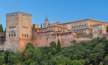 İspanya 'nın Granada kentindeki tarihi Alhambra kompleksi, ön planda sıcak günbatımı ışığı ve yeşillikle birlikte antik savunma kuleleri ve sarayları barındırıyor.