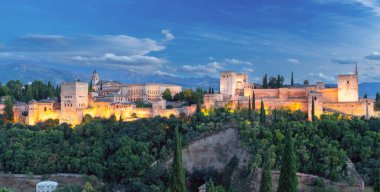 Granada, İspanya 'daki aydınlık Alhambra kompleksinin geniş manzarası, tarihi duvarları ve kuleleri yemyeşil tepeler ve alacakaranlıktaki Sierra Nevada dağlarıyla çevrili.