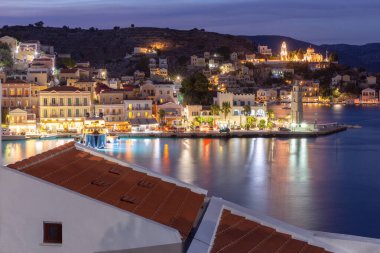 Yunanistan 'ın Symi kentindeki Gialos limanı üzerinde mavi saat manzarası, tepeye tırmanan pastel neoklasik evler ve sakin Akdeniz sularına yansıyan sıcak ışıklar