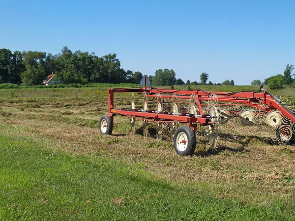 Hay rake sitting in a field of just raked hay with farmhouse in the background. — Stock Photo, Image