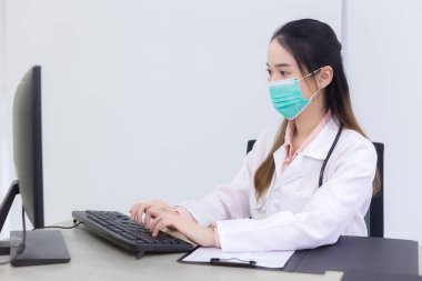 Asyalı kadın doktor hastanede tıbbi maske takarken bilgisayara bilgi kaydetmek için klavyede daktilo kullanıyor..