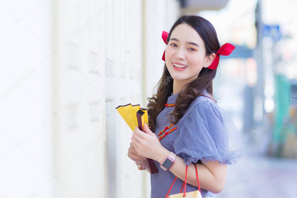 A young Asian woman in a blue-gray cheongsam holds a cloth bag next to a white wooden door with a city background.