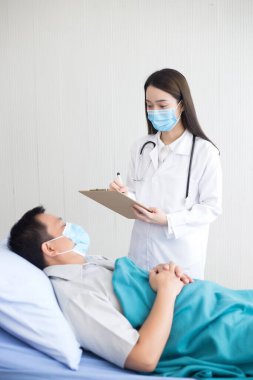Asyalı kadın doktor, Asyalı bir erkek olan ve hastanede yatan hastasının sağlığını teşhis ediyor ve kaydediyor. İkisi de Covid 19 virüsünü korumak için tıbbi maske takıyor..