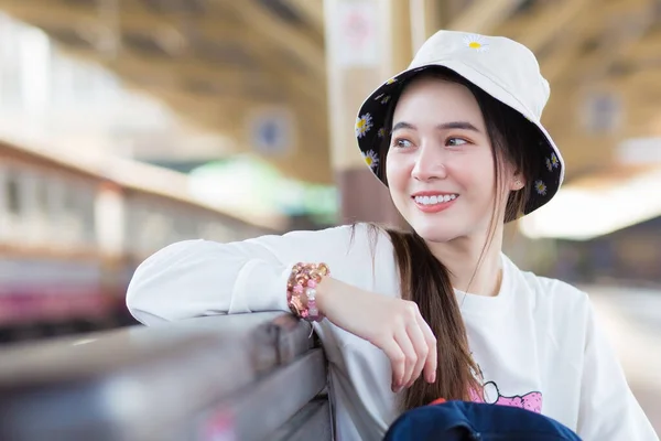 Asian beautiful woman in a long-sleeved white shirt and a hat sits happy smilie in the train station waiting for the train to arrive.