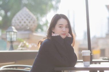 Koyu mavi takım elbiseli Asyalı güzel kadın bir sandalyede oturmuş dışarı bakıyor. Güneşli bir sabahta bir kahve dükkanının önünde..