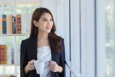 Elinde kahve fincanı tutarken gülümseyen ve pencereden dışarı bakan Asyalı güzel bir ofis kadını. Ev konsepti üzerine çalışıyor..