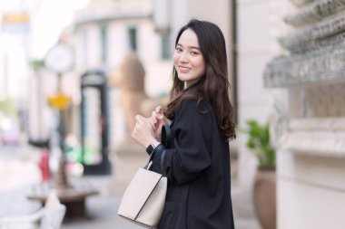 Siyah ceketli, güzel, uzun saçlı bir Asyalı kadının portresi, yürüyüş çantası taşıyor, açık havada mutlu bir şekilde gülümsüyor..