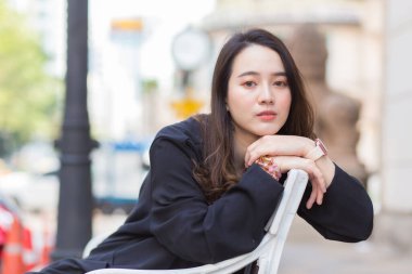 Koyu mavi uzun kollu gömlekli Asyalı güzel bir kadın şehir parkında sandalyede oturuyor..