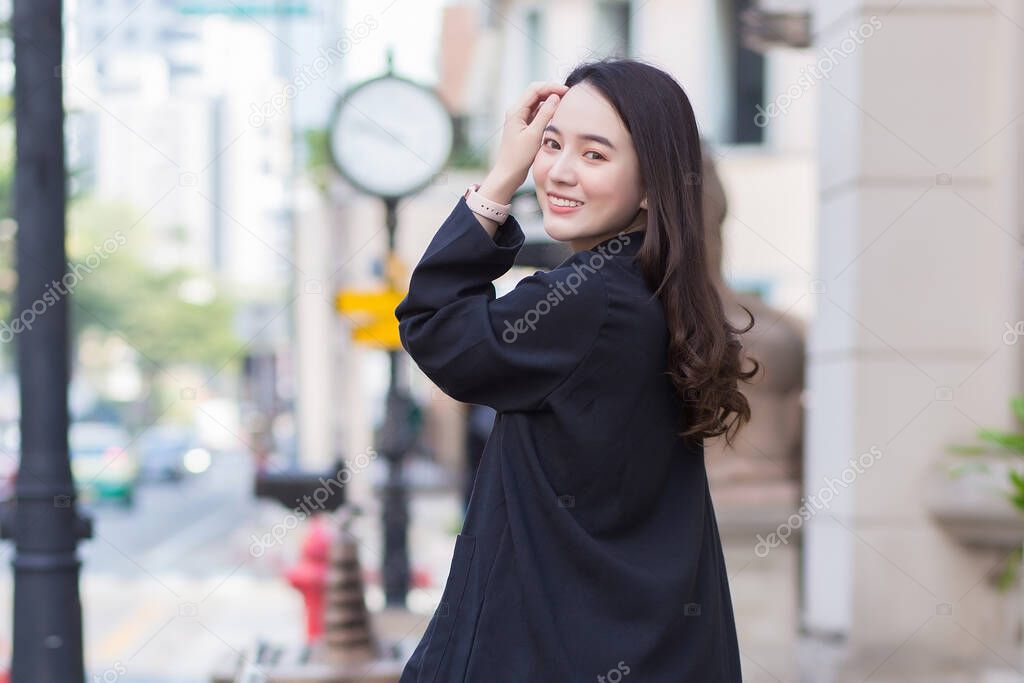 A portrait of a beautiful, long-haired Asian woman in a black coat walking and smiling happy outdoors in the city.