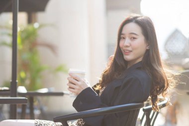 Koyu mavi takım elbiseli Asyalı güzel bayan mutlu bir şekilde bir sandalyeye oturur ve güneşli bir sabahta kafenin önünde kahve fincanı tutar..