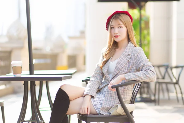Asian beautiful  woman who wears suit and red cap with bronze hair sits on chair in coffee shop while there was a coffee mug on the table on a sunny morning.