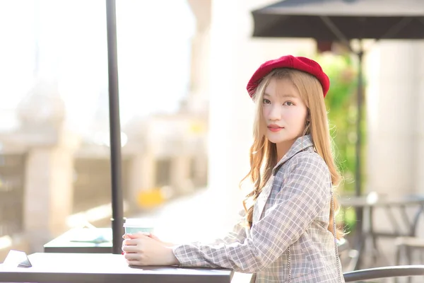 Asian beautiful girl who wears suit and red cap with bronze hair sits on chair in coffee shop while there was a coffee mug on the table on a sunny morning.