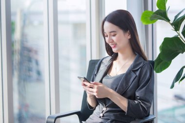 Asyalı işçi kadın gri elbiseler giyer sandalyede oturur ve ofis odasında akıllı telefonu elinde tutar..