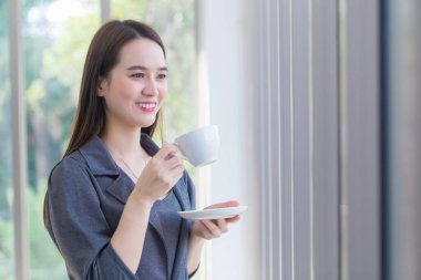 Asyalı çalışan kadın elinde kahve fincanı tutarken bir şey düşünmek için pencereden dışarı bakıyor..