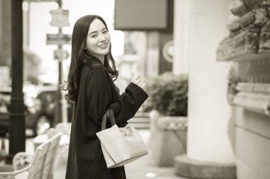 Siyah ceketli, güzel, uzun saçlı bir Asyalı kadının portresi, yürüyüş çantası taşıyor, açık havada mutlu bir şekilde gülümsüyor..