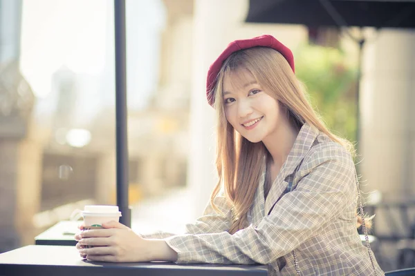 Asian beautiful female who wears suit and red cap with bronze hair sits on chair in coffee shop while there was a coffee mug on the table on a sunny morning.