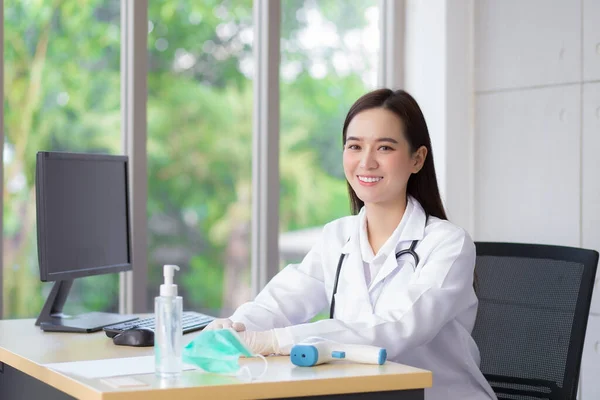 亚洲漂亮的年轻微笑的女医生戴着橡胶手套坐在医院的办公室里 桌上有一张纸 酒精凝胶 温度计和电脑 — 图库照片