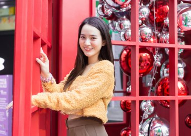Asyalı uzun saçlı, sarı cüppeli güzel bir kadın mutlu bir şekilde kırmızı bir telefon kulübesinin yanında duruyor Noel 'i ve yeni yılı kutlarken.