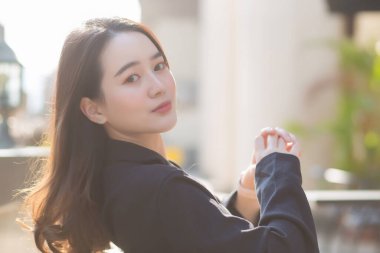 Koyu mavi takım elbiseli Asyalı güzel bir kadın sandalyede oturuyor ve mutlu bir şekilde gülümsüyor. Güneşli bir sabahta kahve dükkanının önünde.