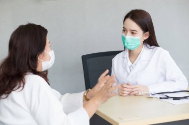Asyalı bir kadın hasta, hastanede enfeksiyonu önlemek için maske takan klinikteki profesyonel bir kadın doktora durumunu özenle açıklıyor..