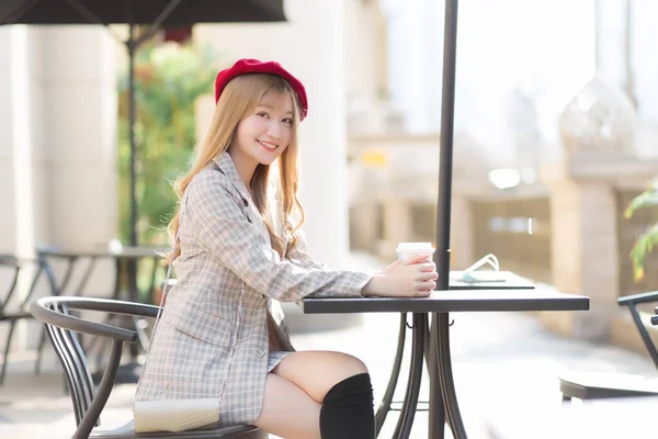Asian beautiful woman who wears suit and red cap with bronze hair sits on chair in coffee shop while there was a coffee mug on the table on a sunny morning.