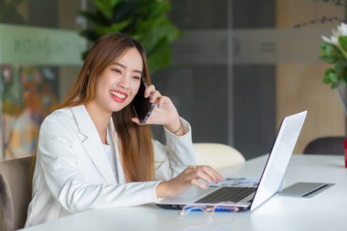 Asyalı güzel, çalışan bir kadın birisiyle konuşuyor. Çalışma odasında ya da ofisinde laptopuyla çalışırken mutlu bir şekilde konuşuyor ve gülümsüyor..