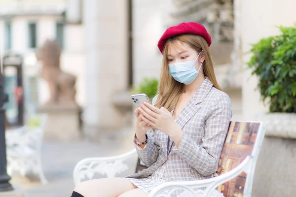 身穿西服 头戴红帽的亚洲女性一边手握智能手机一边坐在公园的长椅上聊天 一边还戴着医疗面罩 污染Pm2 新的保护正常和验尸动物的生活方式理念 — 图库照片