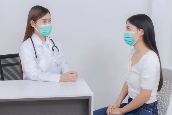 アジアの女性医師は病院の少女患者の症状をチェックするために話している 彼らは両方とも呼吸器系の微生物病原体を保護するために医療面マスクを着用しています — ストック写真
