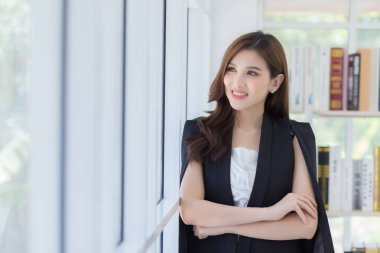 Ev kavramından çalış. Asyalı genç ve güzel bir ofis çalışanı kollarını kavuşturup ofiste bir şeye mutlu bir şekilde gülümsüyor..