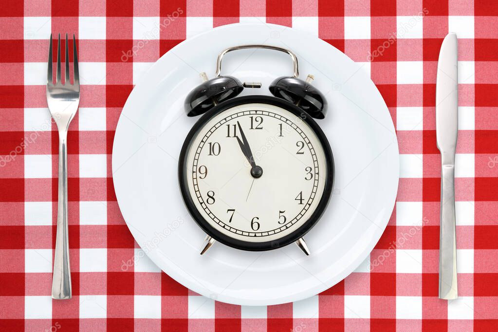 Time to Eating or Intermittent fasting concept - flat lay composition with alarm clock, plate and utensils on a red checked tablecloth.
