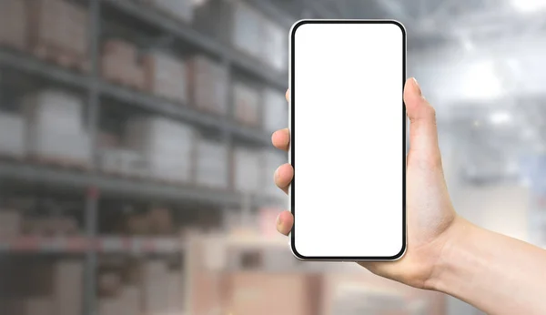 Modern warehouse management template - female hand holding frameless smartphone with blank white screen on blurred background of delivery company interior.