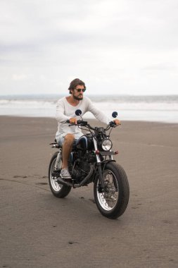Sahilde motorsiklet süren genç bir adam, açık hava portresi, güneş gözlüğü, şort, spor ayakkabı ve tişört, birlikte seyahat, okyanus, deniz...