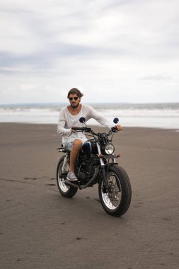 Sahilde motorsiklet süren genç bir adam, açık hava portresi, güneş gözlüğü, şort, spor ayakkabı ve tişört, birlikte seyahat, okyanus, deniz...