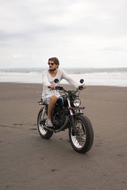 Sahilde motorsiklet süren genç bir adam, açık hava portresi, güneş gözlüğü, şort, spor ayakkabı ve tişört, birlikte seyahat, okyanus, deniz...