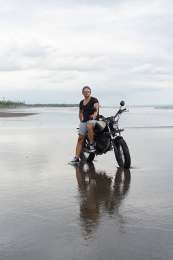 Sahilde motorsiklet süren genç bir adam, açık hava portresi, güneş gözlüğü, şort, spor ayakkabı ve tişört, birlikte seyahat, okyanus, deniz...