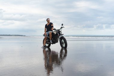 Sahilde motorsiklet süren genç bir adam, açık hava portresi, güneş gözlüğü, şort, spor ayakkabı ve tişört, birlikte seyahat, okyanus, deniz...
