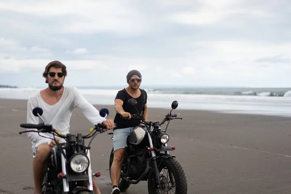Two young men friends ride a retro motorcycle on the beach, outdoors portrait, posing, in sunglasses, shorts, sneakers and t-shirts, travel together, ocean, sea