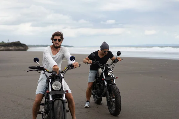 Two young men friends ride a retro motorcycle on the beach, outdoors portrait, posing, in sunglasses, shorts, sneakers and t-shirts, travel together, ocean, sea