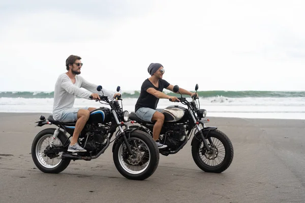 Two young men friends ride a retro motorcycle on the beach, outdoors portrait, posing, in sunglasses, shorts, sneakers and t-shirts, travel together, ocean, sea