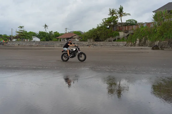 Young man riding a retro motorcycle on the beach, outdoors portrait, posing, in sunglasses, shorts, sneakers and t-shirts, travel together, ocean, sea