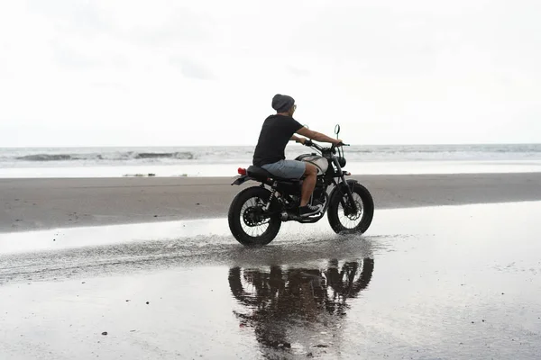 Young man riding a retro motorcycle on the beach, outdoors portrait, posing, in sunglasses, shorts, sneakers and t-shirts, travel together, ocean, sea