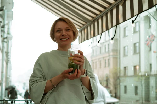 オープンテラスで雨の中の若い女性が大きなガラスからお茶を飲む — ストック写真